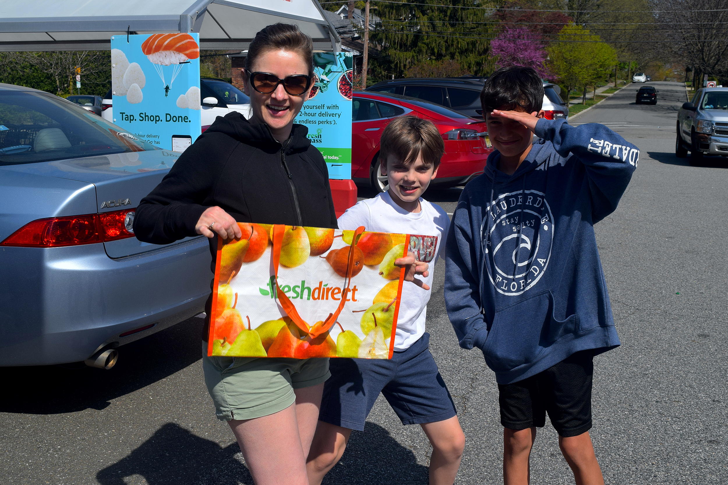 A woman holds a reusable shopping bag in a parking lot, next to two children