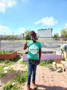 A woman in a green t-shirt and jeans gives a peace sign in front of raised garden beds