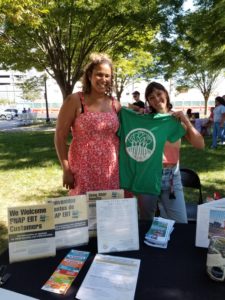 Two woman stand behind a table covered in informational material; one woman holds up a green t-shirt