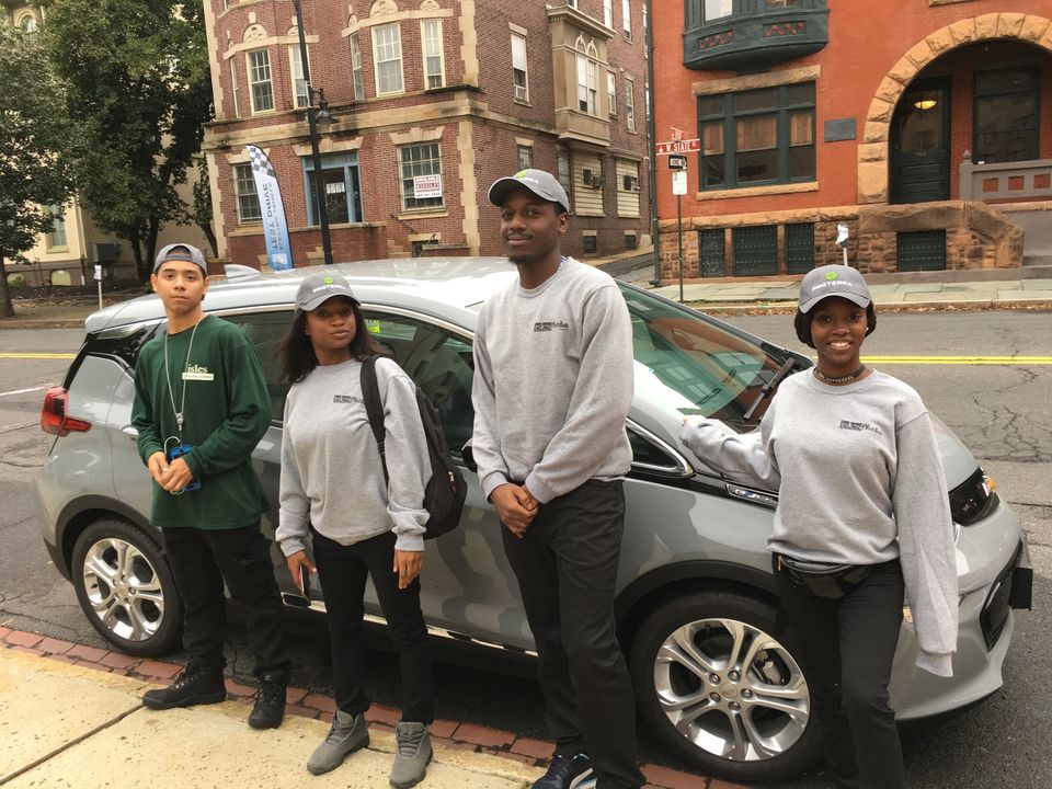 Four smiling people stand in front of a vehicle on a city street