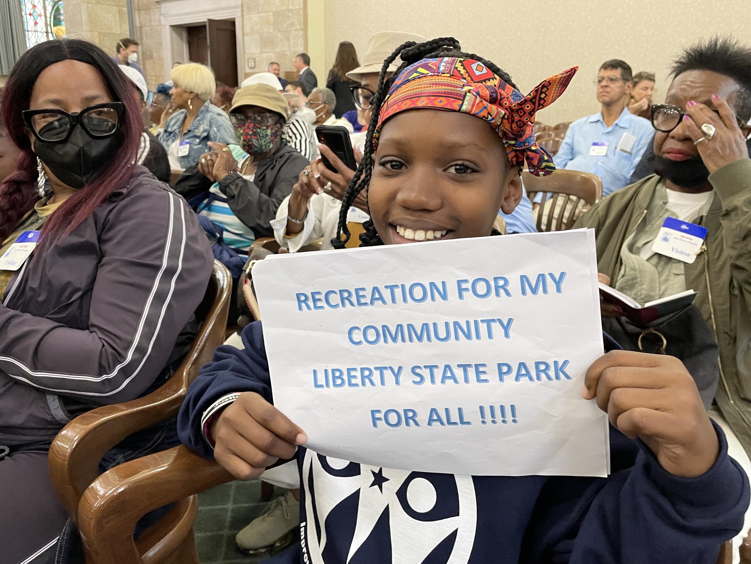 A child sitting in an audience of people holds a sign stating "RECREATION FOR MY COMMUNITY LIBERTY STATE PARK FOR ALL!!!!"