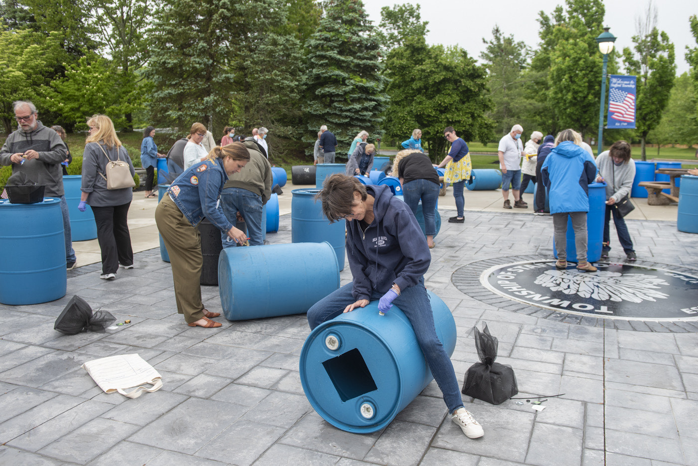 In the foreground, a person sits astride a large blue barrel; people cluster around other barrels in the background