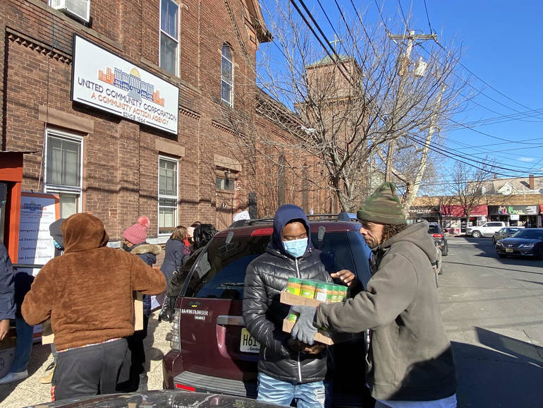 A few people in cold-weather gear carry boxes of food outside a brick building with the sign "United Community Corporation"