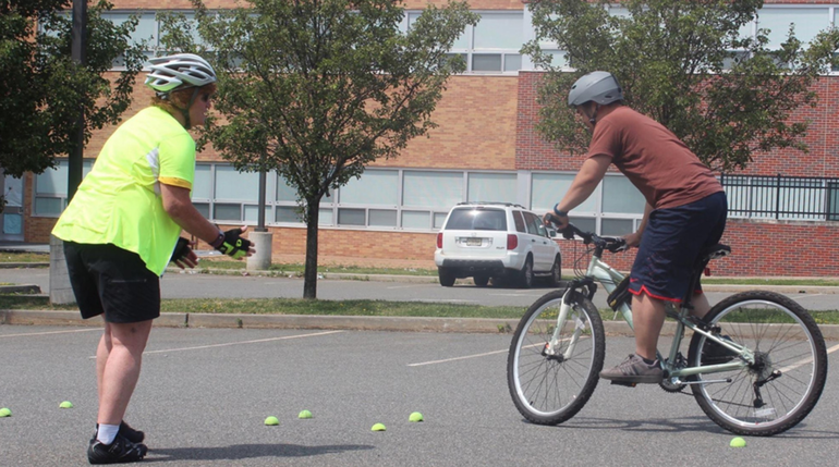 On the left, a person stands in a bike helmet, watching a person on a bike in what appears to be a training course set up in a parking lot