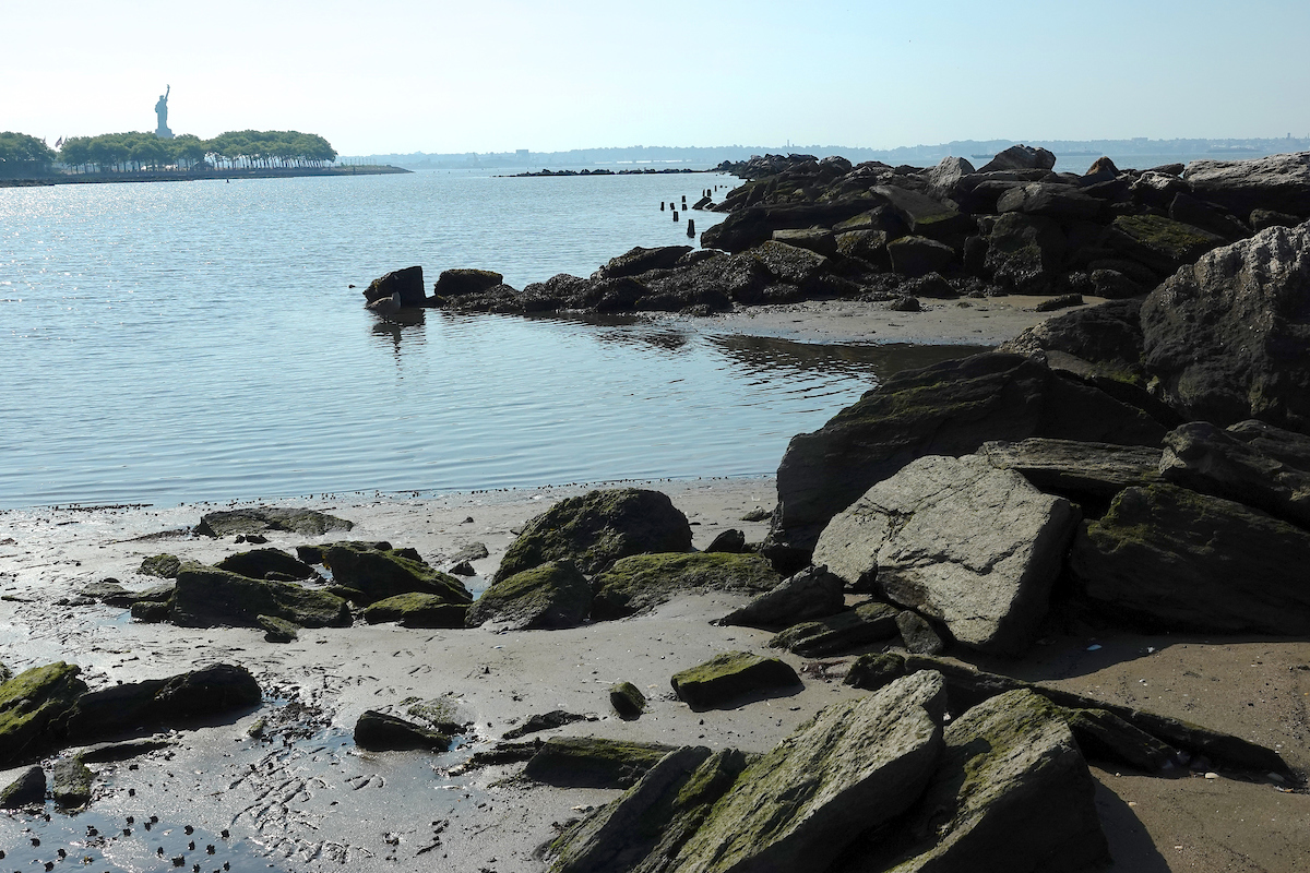 A beach with big rocks foregrounds water and the Statue of Liberty tiny in the background
