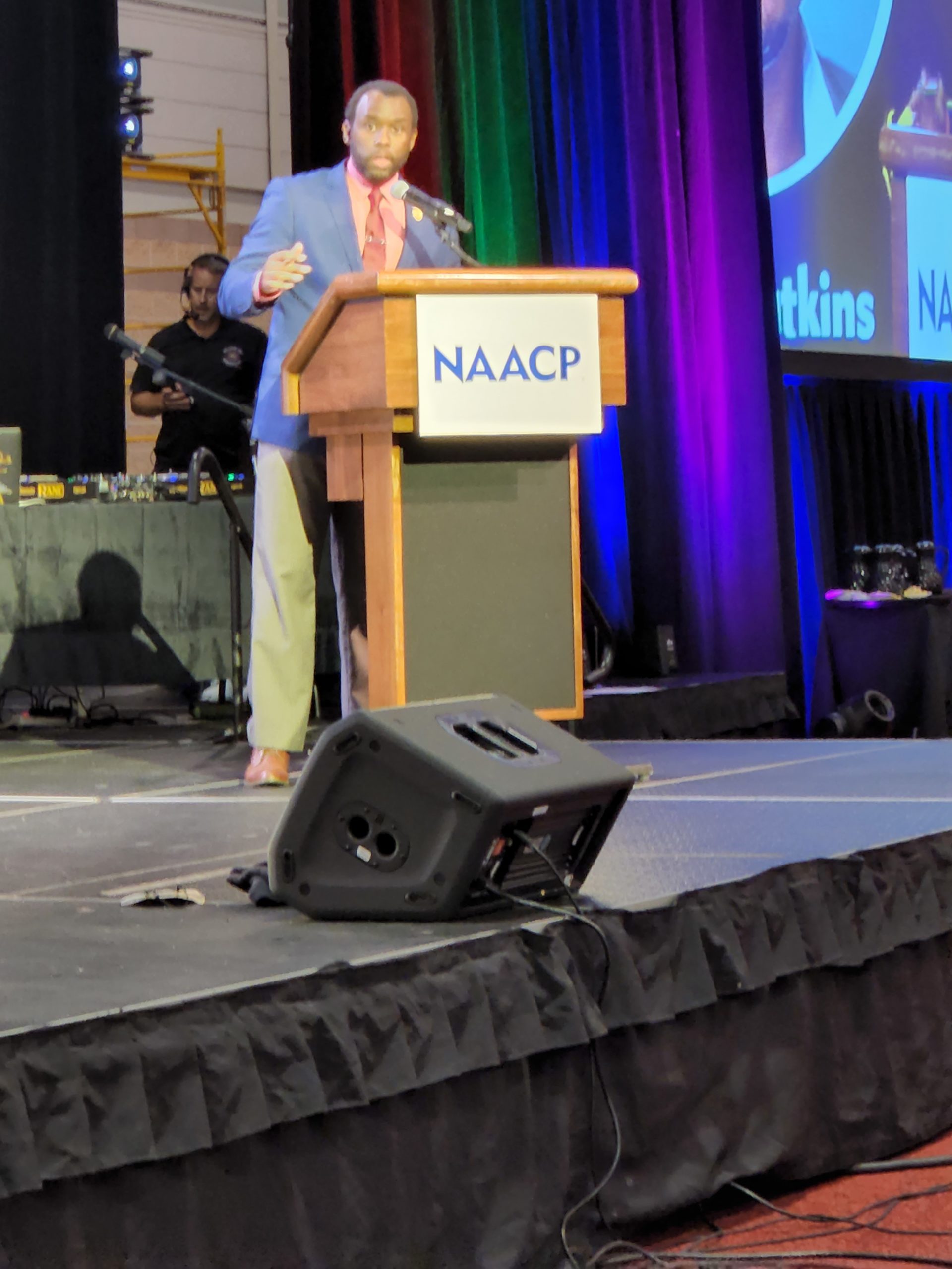 A man in a jacket and tie speaks from a podium with an "NAACP" sign