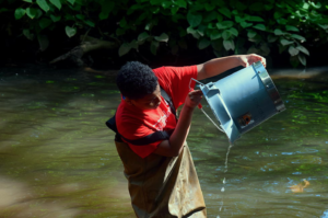 A person in waders stands in water and empties a bucket