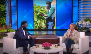 Two people face each other in white arm chairs on a television talk show set, with a picture of one of them gardening displayed on a screen in the background.