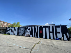 Asphalt leads up to a large mural with a person'a face and the words "BE KIND 2 ONE ANOTHER"