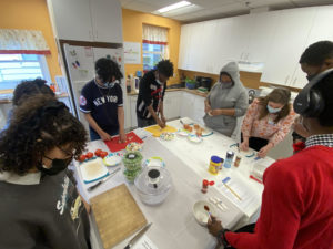 A group of about 9 young people in face masks surround a large table, upon which they are handing food items