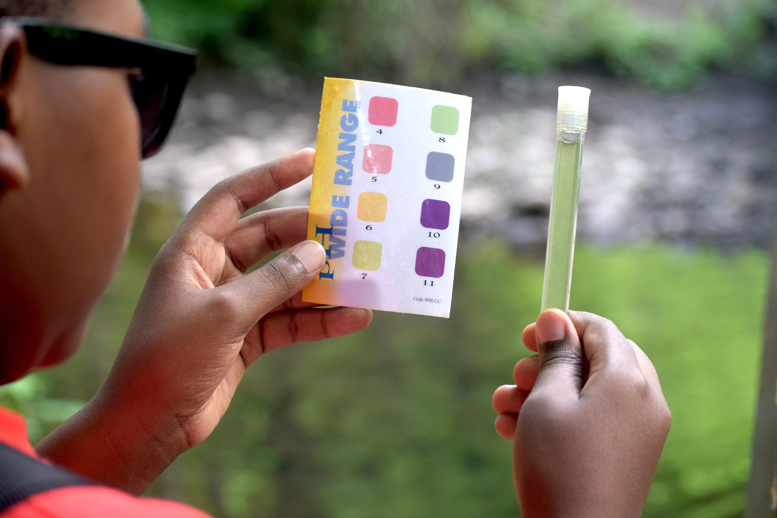 A person looks at a vial of liquid and a small sheet showing colors with the words "pH WIDE RANGE"