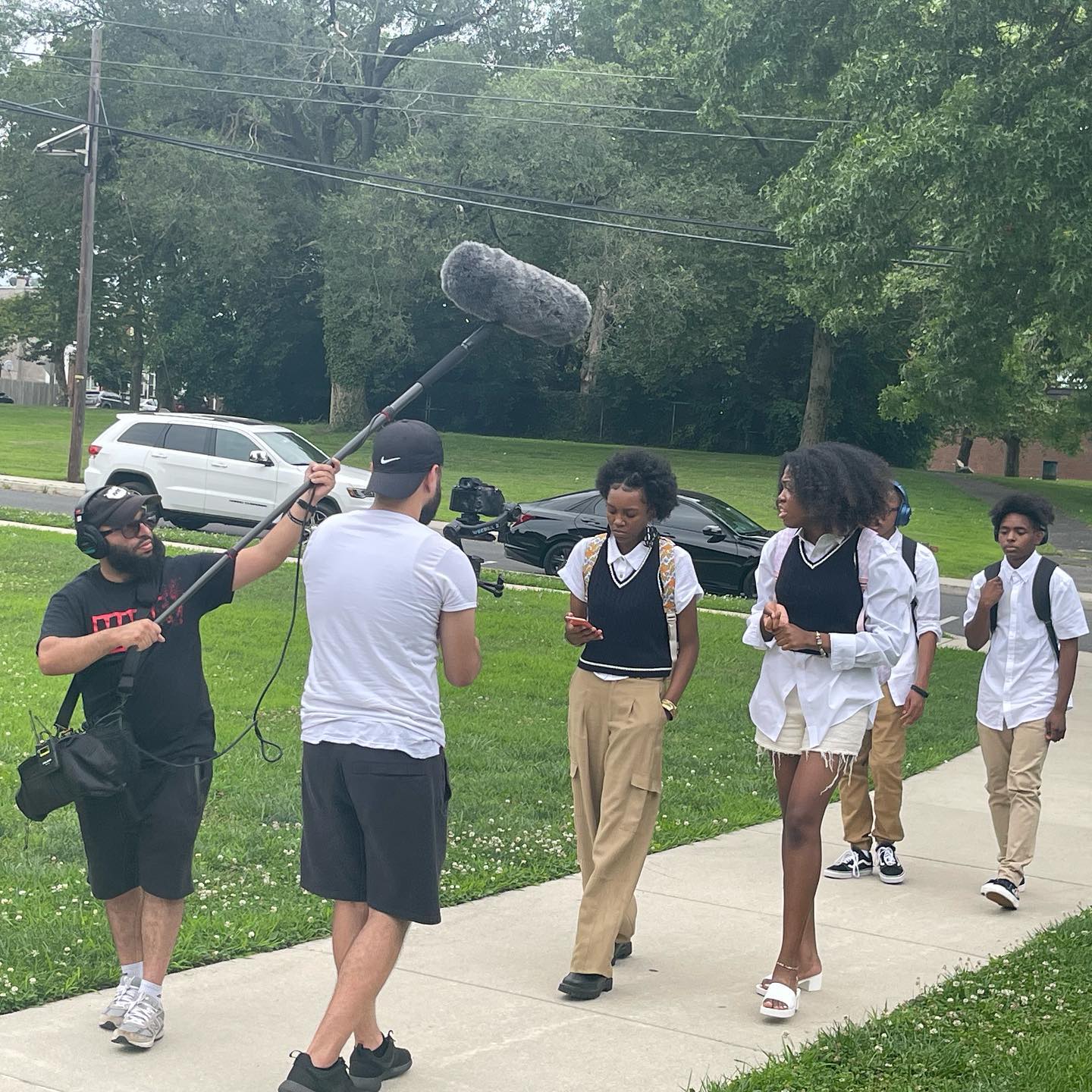 Four young people walk up a sidewalk, recorded by a man holding a video camera and another man holding a microphone boom
