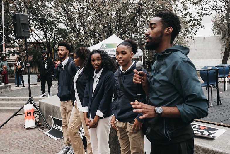 Five young people stand in a line, one of them speaking into a microphone
