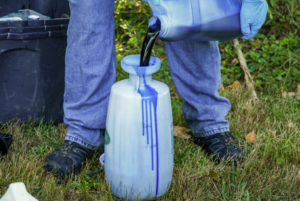 A blue liquid is poured from one container to another by a person standing in grass