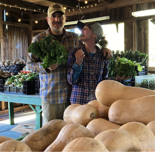 A man and woman, each holding a green leafy vegetable, stand together behind a pile of large squashes