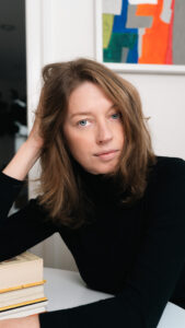 A light skinned woman with light brown hair, wears a dark sweater and leans on a table on her right elbow