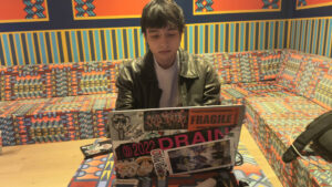 A young man sits in front of his laptop. Various objects are on the table.