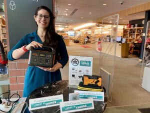 A light skin woman wearing glasses holds with both hands, a device containing outlets for USB, electric and other ports. In the background are rose of books and assign that says library of things