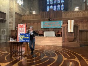 A woman wearing glasses with short, curly hair stands at a lectern. On it is a sign that says make polluters pay. Another sign in the background reads no more fossil fuels.